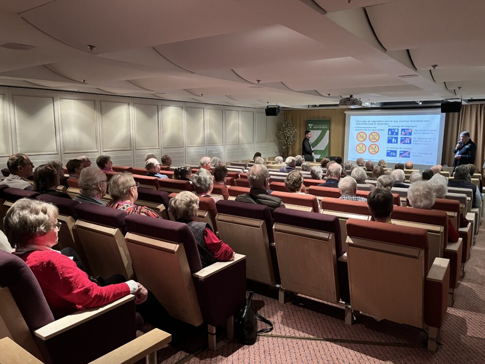 En grupp äldre vuxna sitter i ett litet auditorium och tittar på en presentation. Två presentatörer står längst fram vid en skärm som visar diabilder med olika symboler och text.