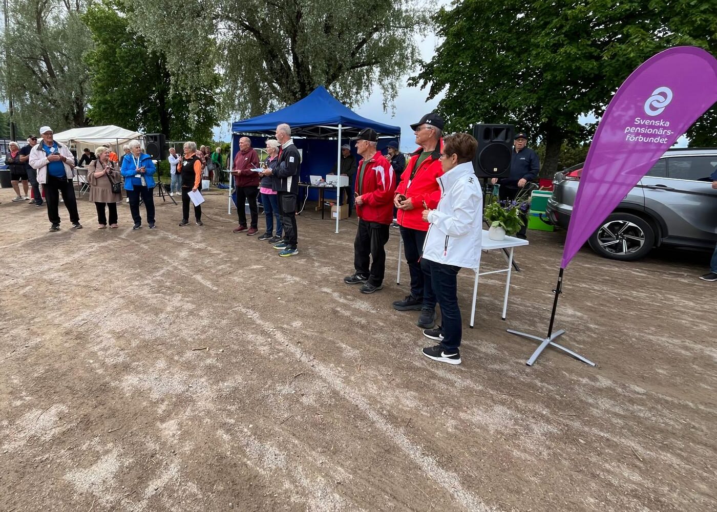 En grupp människor står på en grusplan nära ett bord med troféer, en lila banderoll med texten ”Svenska Pensionärers Förbund” och blå tält. Vissa verkar ta emot utmärkelser medan andra tittar på. I bakgrunden syns träd och bilar.