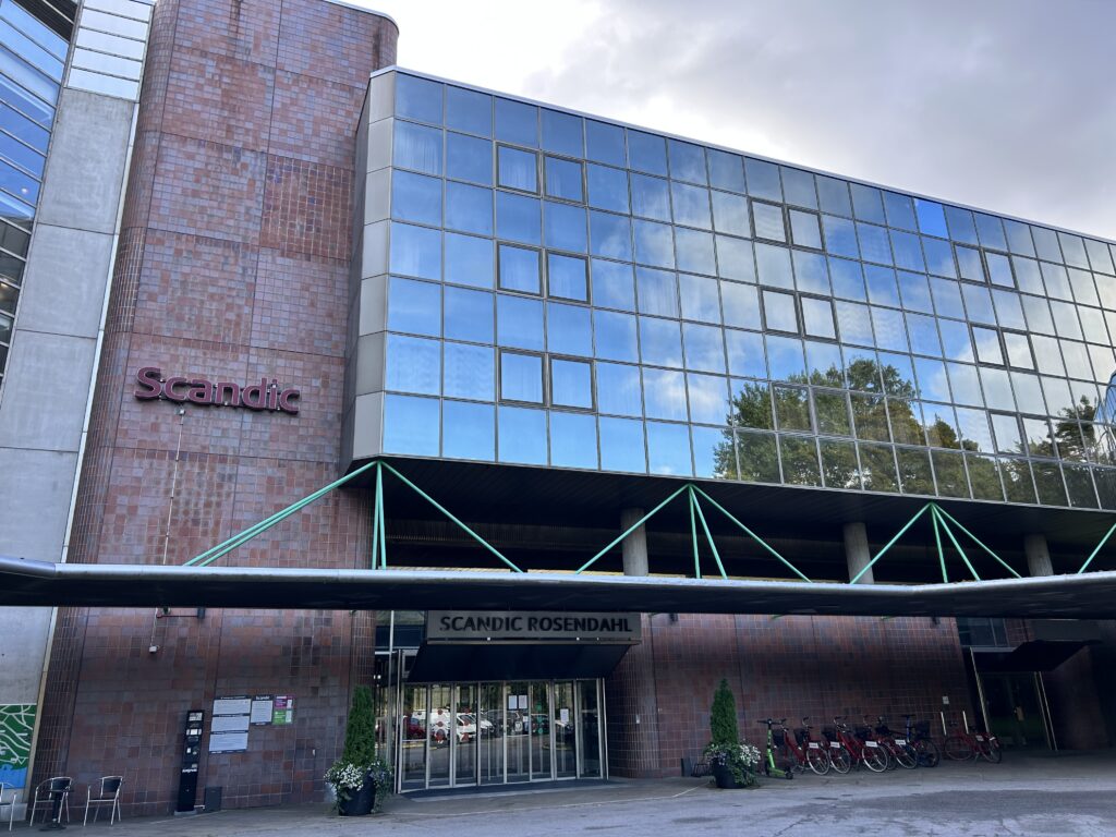 Bilden visar utsidan av hotellet Scandic Rosendahl, med stora glasfönster, en tegelfasad och en överbyggd entré. Flera cyklar står parkerade nära entrén.
