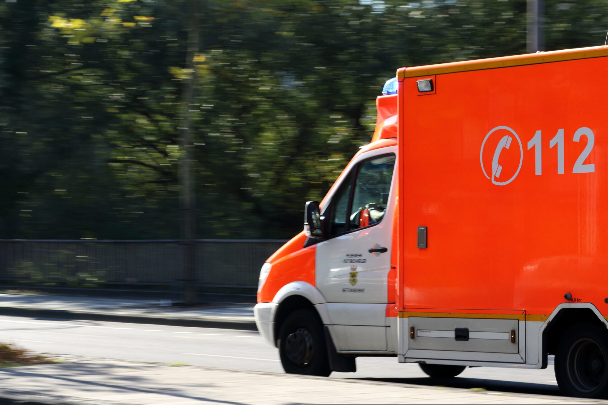 En knallorange ambulans med numret 112 och en telefonsymbol kör snabbt längs en stadsgata, med en rörelseoskärpa i bakgrunden där träd och ett metallräcke syns, vilket ger intryck av brådska eller en räddningsinsats.