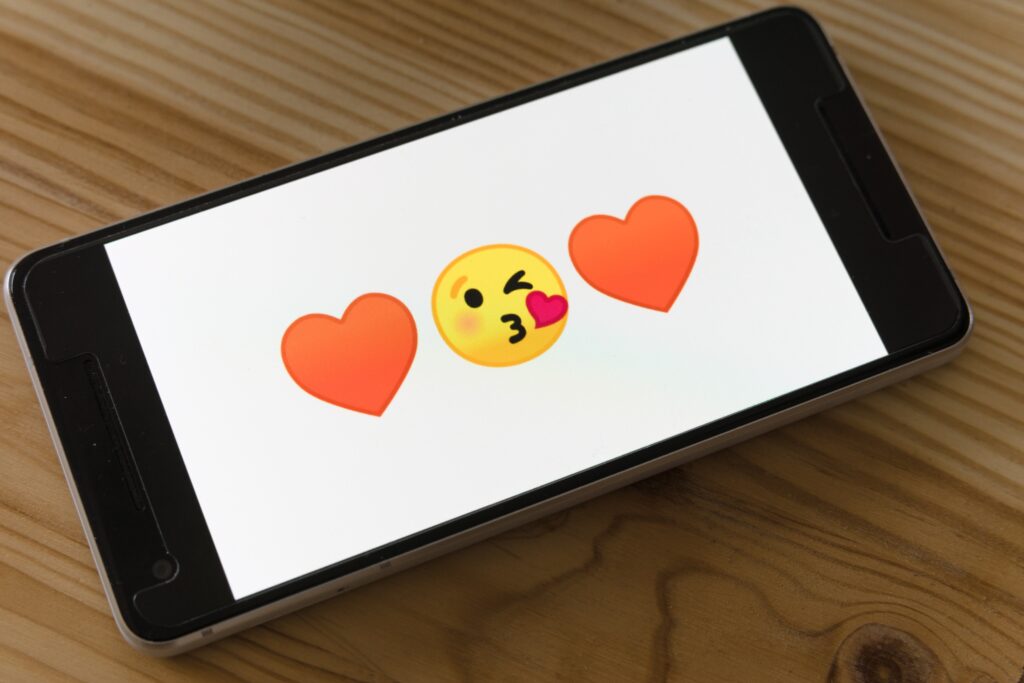 På skärmen på en smartphone som ligger på en träyta visas två röda hjärt-emojis och en emoji för ett blinkande ansikte som blåser en kyss.