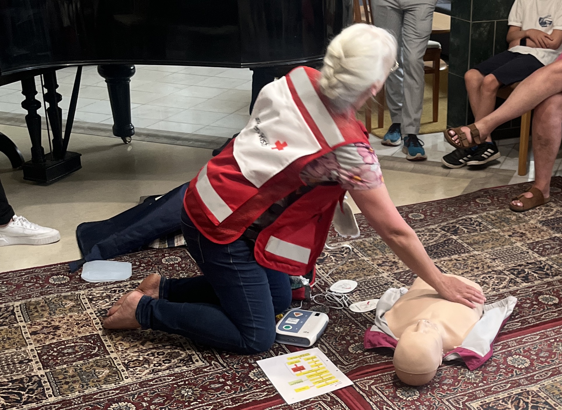 En person i en röd och vit väst visar hur man utför hjärt-lungräddning på en övningsdocka i ett rum med en mönstrad matta. Flera personer sitter runt omkring och tittar på demonstrationen. Material för hjärt-lungräddningsutbildning syns på golvet.