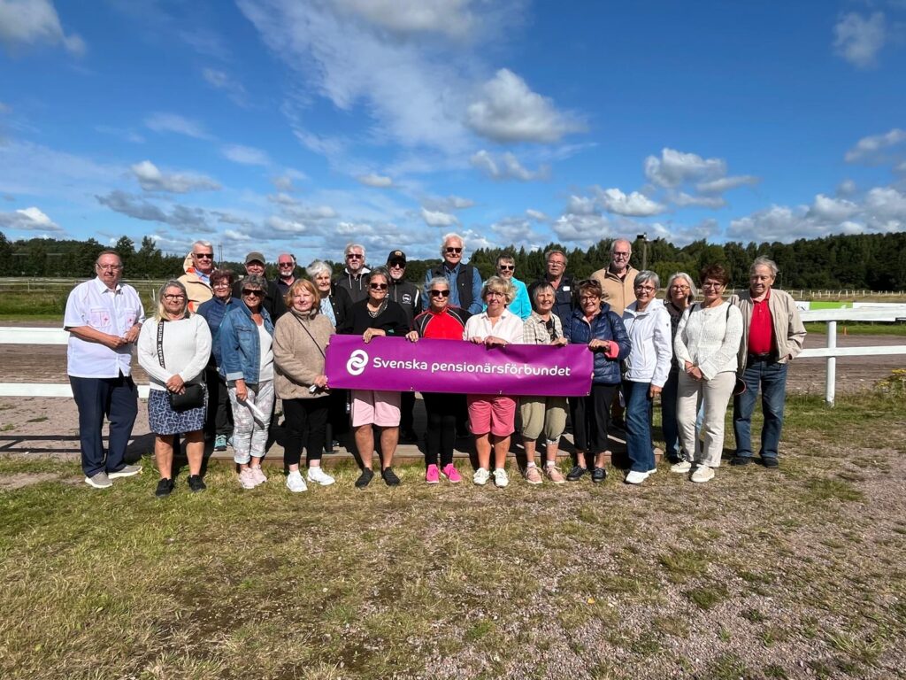 En grupp vuxna poserar utomhus på gräset under en blå himmel och håller i en lila banderoll med texten ”Svenska pensionärsförbundet”. I bakgrunden syns ett vitt staket och träd.