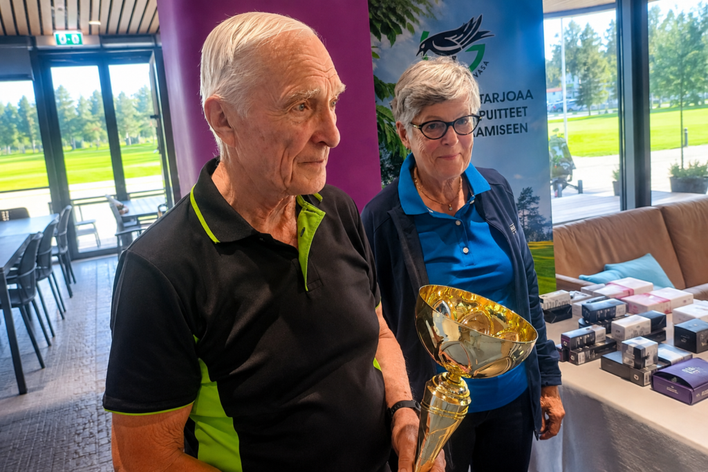 En äldre man i en svart och grön skjorta håller i en guldpokal. En äldre kvinna i en blå jacka står bredvid honom. De befinner sig inomhus nära bord med små lådor på, och genom fönstren syns träd och gräs utanför.