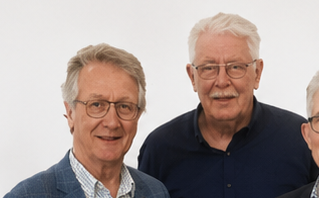 Två äldre män med grått hår och glasögon står tillsammans framför en enfärgad ljus bakgrund. Båda tittar in i kameran och ler lätt. Endast överkropparna och en del av en tredje person syns.
